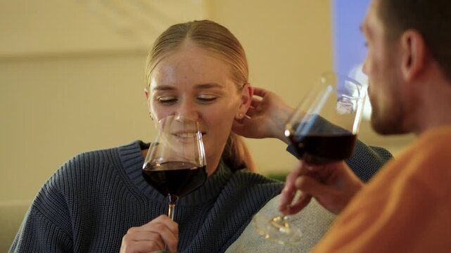 Blushing smiling woman sips red wine during cozy evening date on comfortable couch. Romantic couple shares tender conversation, romance and first meeting, exchanging loving glances, tender couple