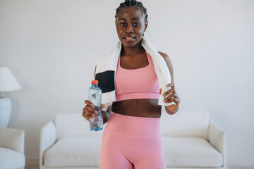 Towel on neck and bottle of water in hand. Woman is doing yoga at home