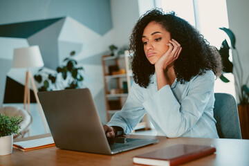 Young woman with curly hair seated at a desk with a laptop, showing contemplative expression in a cozy home office setting