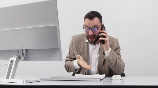 Angry businessman shouting and gesticulating while talking on cell phone in front of computer display on light background. The concept of rage, aggression and discontent.