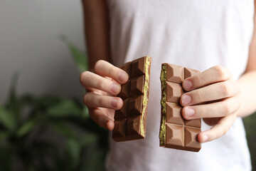 Front view of hands breaking a pistachio-filled chocolate bar. Person in white tank top, natural soft light, blurred green plant background. Focus on texture and real moment. High quality photo