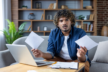 A shocked man stares at documents while sitting on the sofa with a laptop. Financial difficulties...