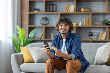 Young man with curly hair is sitting on the sofa at home shopping online, using a phone and a credit card for the payment transaction.
