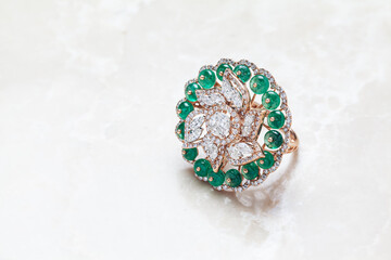 Close-up of a diamond ring having many diamonds and emerald stones over a white marble background.