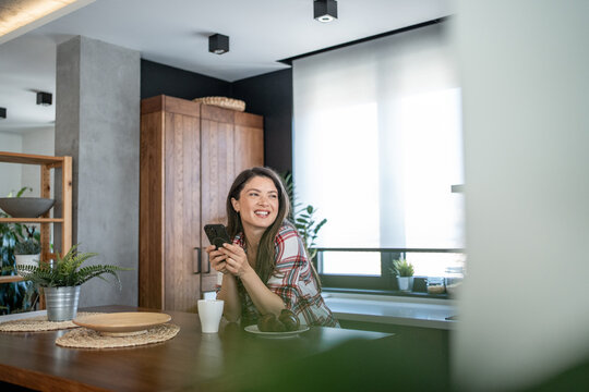 Smiling woman using smartphone and enjoying coffee break in modern kitchen - Powered by Adobe