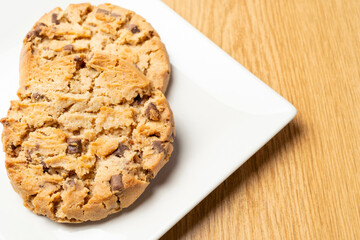 Two freshly baked chocolate chip cookies sit on a white square plate, ready to be enjoyed, on a wooden table