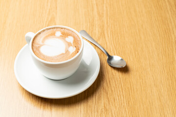 A cup of hot moccacino with creamy latte art, served on a white saucer with a spoon, placed on a wooden table, creating a warm and inviting scene