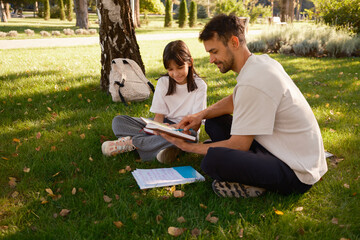 A father and daughter share a moment reading a book outdoors on a sunny day, enjoying the peaceful park environment and quality time together. The image captures family bonding and the joy of learning