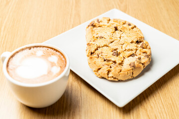 Two chocolate chip cookies on a white plate beside a cup of cappuccino with latte art, placed on a wooden table, creating a cozy and inviting atmosphere.