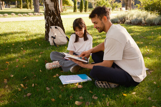 A father and daughter share a moment reading a book outdoors on a sunny day, enjoying the peaceful park environment and quality time together. The image captures family bonding and the joy of learning
