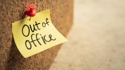 Out of office note on yellow sticky pad pinned to corkboard, indicating absence from work, office communication concept