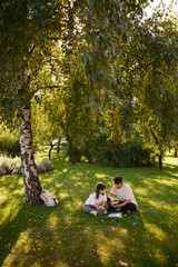 A father and daughter sit on the grass in a park, surrounded by nature. They are engaged in...