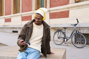 Young man wearing headphones using phone and listening to music, sitting in the city with his bicycle behind him