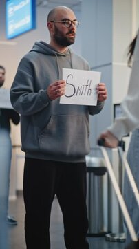 International Airport Terminal: Airport Greeter Holding Name Sign, Meeting Passengers From Plane Flight. Diverse People Walking with Luggage. Business Transfer or Transportation Service. Vertical Shot