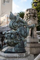 Two weathered stone Komainu, guardian lion dogs, stand on tiered granite bases at Namba Yasaka Jinja Shrine in Osaka, Japan. One is bronze, the other light grey. 