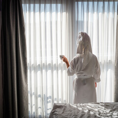 Beautiful young woman in a white bathrobe with a towel on her head stands near the window.