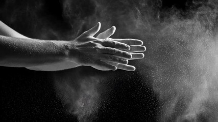 Black and white shot of hands clapping together, creating a cloud of white powder (talc or chalk) against a dark background, conveying impact, energy, celebration or performance preparation