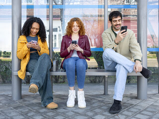 Three diverse young people using smartphones while waiting at a bus stop