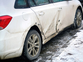 White car with extensive dents, scratches and chipped paint on its side, accident damage. Side...
