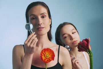 Women holding flowers and glass objects in artistic pose against blue background