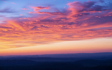 A vibrant sky ablaze with colors at dusk or dawn, showcasing hues of pink, orange, and blue.  Clouds are painted with light above a dark silhouette of distant mountains at the horizon.