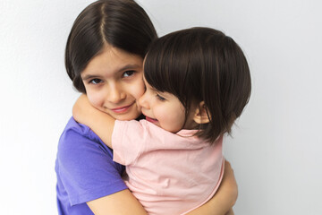 Two young sisters hugging each other warmly, older girl smiling gently while holding her little sister close against plain white wall, close up portrait