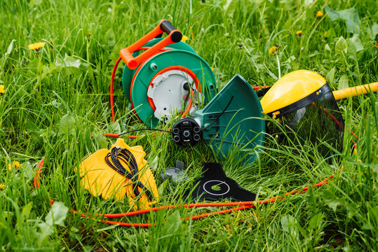 Equipment for mowing grass. Electric trimmer scythe, garden reel with red wire, safety helmet, shield, gloves, metal knife blade, grass line. Green grass background.