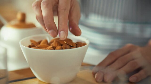 A person carefully picks almonds from a white bowl in a well-lit kitchen. The activity takes place during a sunny day, focusing on healthy snacking and food preparation