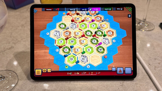 A digital game of Catan displayed on a tablet during a casual board game night, showing the hexagonal resource tiles, settlements, and roads in mid-play, surrounded by drinks and Apple Pencil.