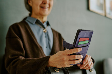 Senior woman holding a smartphone while smiling, captured in indoor setting with a contemporary feel, she wearing brown sweater over blue shirt, exuding warmth and positivity