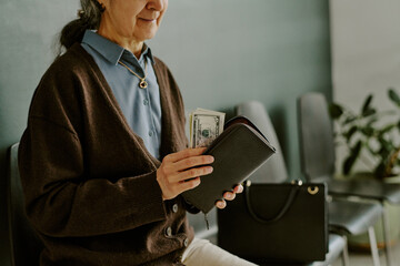 Senior woman holding open leather wallet with dollars in it while sitting in modern waiting area