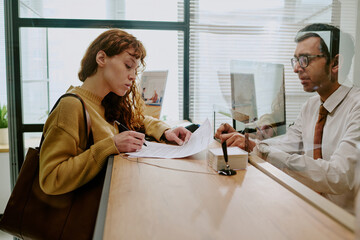 Young woman concentrating while signing document at bank office counter with professional assisting, busy office environment with natural light illuminating the setting