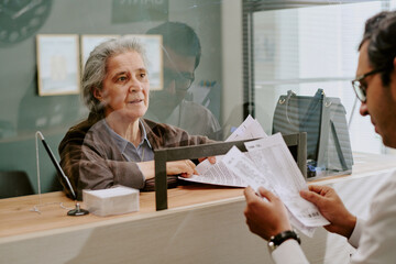 Senior woman behind reception desk assisting a man with important documents. Interaction happening...