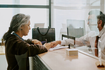 Elderly woman discussing and signing documents at modern office counter while young man assists...