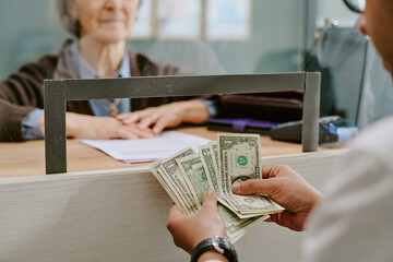 Senior bank clerk handling financial transaction for customer across glass divider in bank branch...