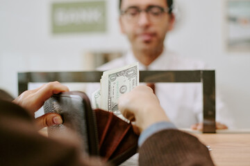 Person holding wallet while giving cash to bank teller standing behind counter, executing banking...