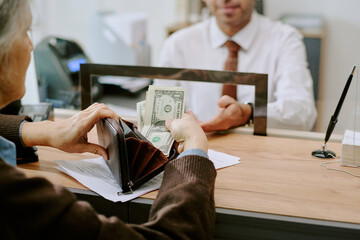 Senior person handing money to bank employee across counter in professional setting, financial...