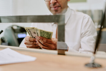 Bank employee counting cash bills at counter behind transparent glass partition in workplace...