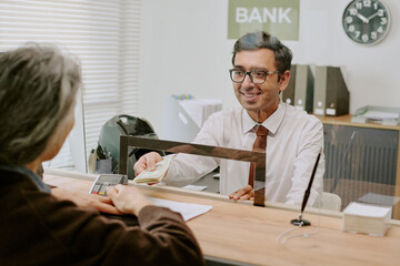 Bank teller wearing glasses and a white shirt smiling while assisting customer by handing currency...