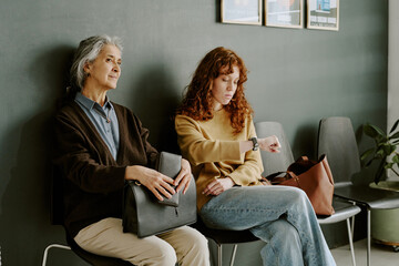 Two women sitting in waiting area of office, one with folder and business attire, other wearing casual clothes