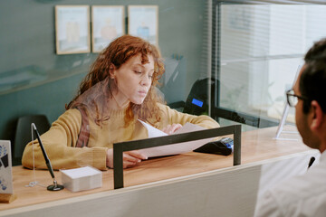 Young woman with curly hair sitting at reception desk while handling paperwork, interacting with...