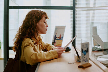 Woman with curly hair reading paper document at reception desk in modern bank office setting, with...