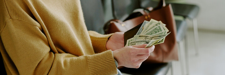 Person in yellow sweater counting cash while holding a wallet, leather handbag on a chair, suggesting surroundings of a public area or office setting
