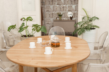 Wooden dining table set with a teapot and multiple cups invites a cozy gathering. Surrounded by transparent chairs and potted plants, the room exudes warmth and comfort.