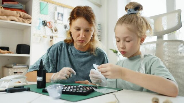 Mother and daughter enjoy creative time making resin jewelry at home. Child learns to mix colors and pour resin into molds, creating handmade accessories while bonding through fun and artistic hobby.