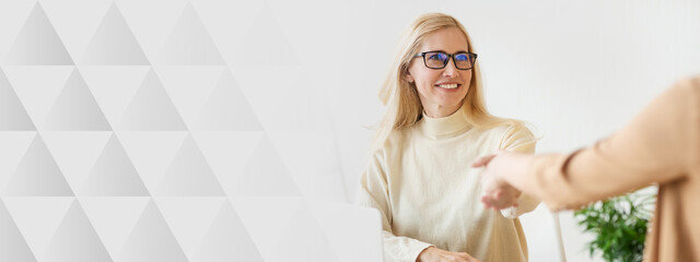 A woman wearing glasses sits at a desk, smiling warmly as she reaches out her hand for a handshake....