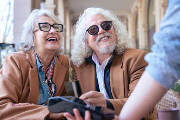 Joyful caucasian senior couple at an outdoor cafe making a card payment. Both wearing trendy outfits and sunglasses. Ideal for retirement, finance, lifestyle, and senior couple relationship concepts. 