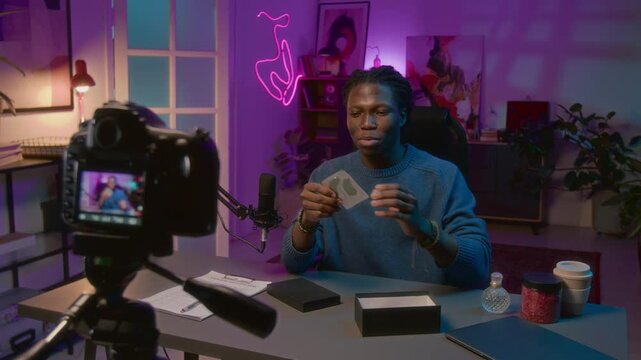 Medium shot of digital video camera on tripod filming cheerful African American male influencer opening box with branded cosmetics on live stream and promoting eye patches to subscribers