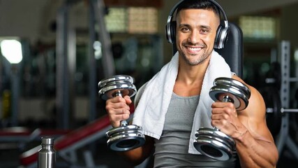 A man smiles confidently while lifting dumbbells in a gym setting. He wears headphones, a towel around his neck, and has a water bottle beside him as he focuses on his workout