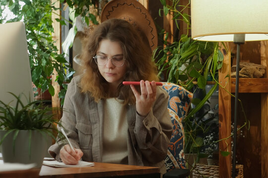 Female employee talking on mobile phone using speakerphone taking notes sitting at desk. 